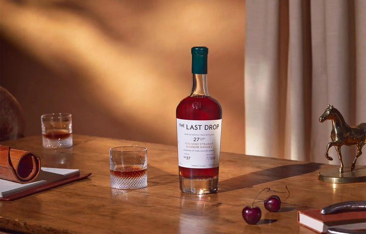 A bottle of The Last Drop Release No. 37 Buffalo Trace 27 Year Old Kentucky Straight Bourbon Whiskey 700ml, a rare expression from Buffalo Trace, sits elegantly on a wooden table alongside two glasses of whiskey, a pair of cherries, a notebook, a pen holder, and a small horse statue in the background—evoking the rich heritage synonymous with Buffalo Trace Distillery.