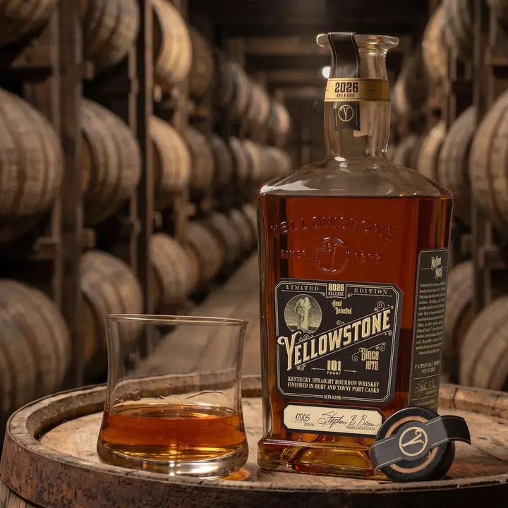 A bottle of Yellowstone 2026 Limited Edition Bourbon Finished in Ruby and Tawny Port Casks, a glass of Kentucky straight bourbon, and a cap sit on a barrel in a dimly lit aging room lined with wooden barrels.