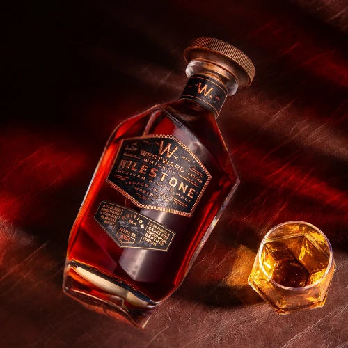 A bottle of Westward Whiskey Milestone Edition No. 4 American Single Malt (Pre-Order) rests on a brown surface beside a glass filled with the same whiskey, both highlighted by warm, angled lighting.