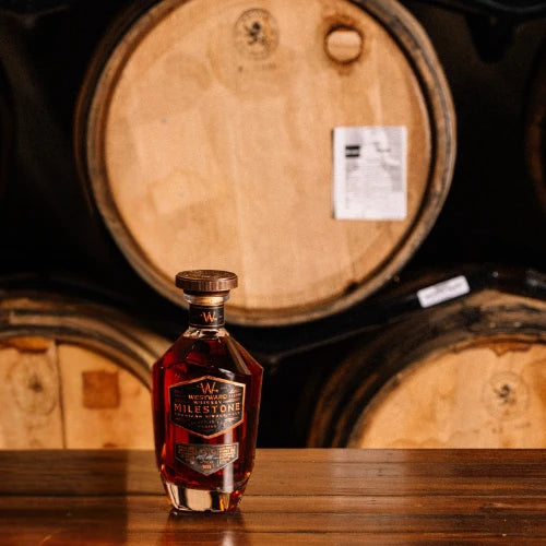 A bottle of Westward Whiskey Milestone Edition No. 4 American Single Malt (Pre-Order) from Westward rests on a wooden surface before towering barrels, softly illuminated to create a warm, inviting atmosphere that showcases the craftsmanship behind this exceptional whiskey.