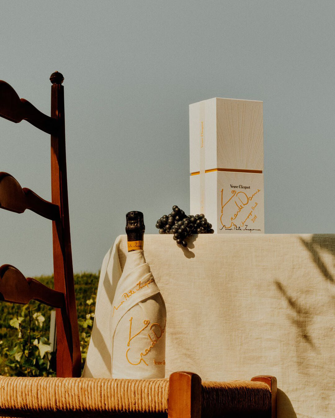 A wooden chair stands by a table with a light cloth, displaying Veuve Clicquot La Grande Dame 2018 Limited Edition by Simon Porte Jacquemus, grapes, and a white gift box outdoors amid lush greenery.