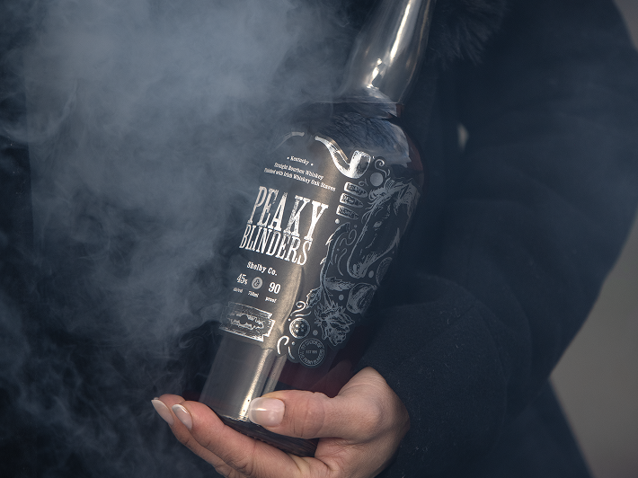 A person in a dark coat holds a shiny Peaky Blinders Irish Whiskey Shelby Company Limited Edition bottle amid swirling smoke, with only their hands and part of the 90 proof Peaky Blinders whiskey bottle visible.