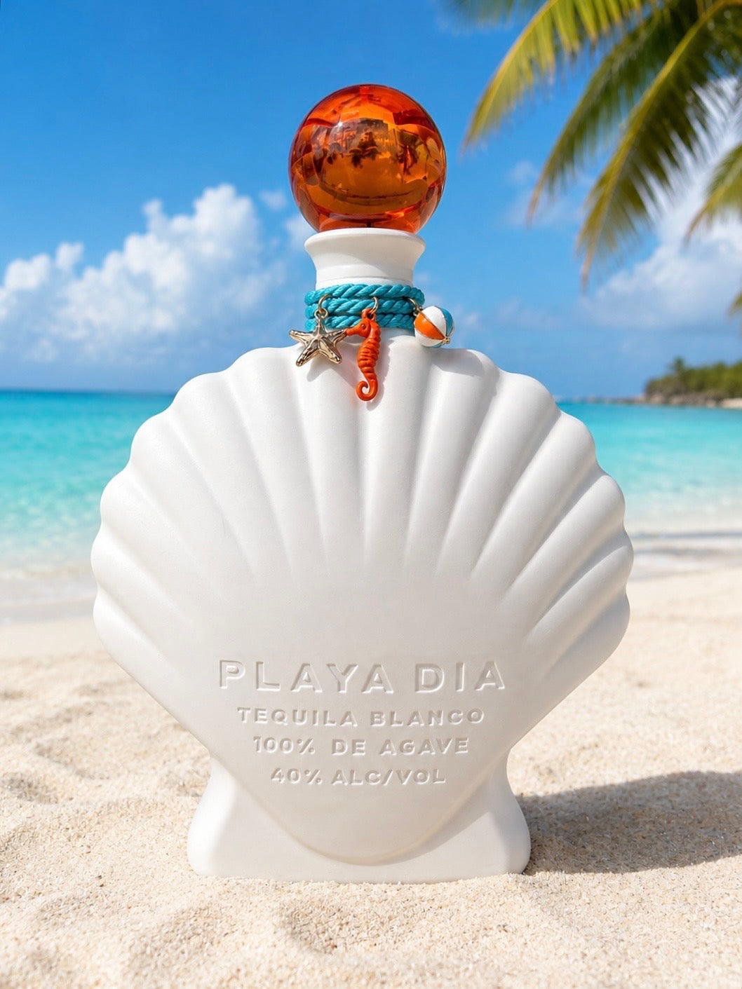 A shell-shaped white bottle of Playa Dia Blanco Tequila rests on a sandy beach with a turquoise sea, palm frond, and blue sky in the background; this 100% Blue Weber agave, additive-free tequila features an orange stopper and is adorned with seashell charms.