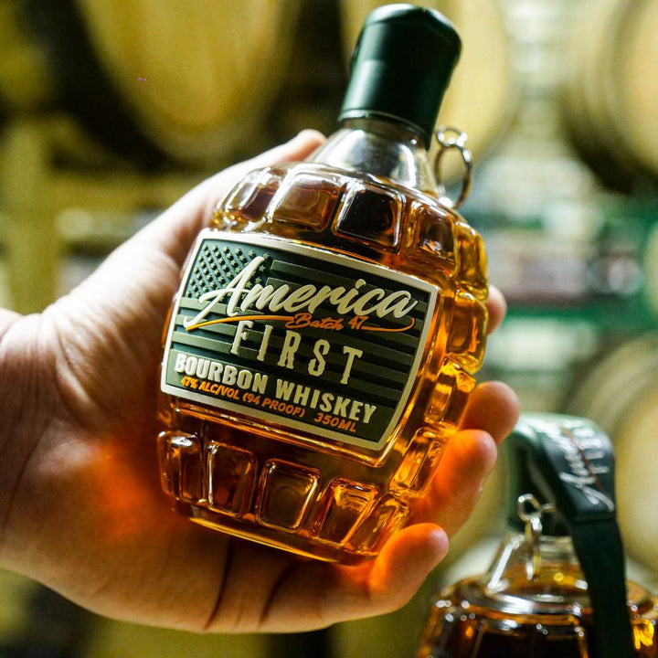 A hand holds an America First Grenade Straight Bourbon Whiskey 2 Pack bottle, shaped like a grenade and labeled 94 proof, 350ml—ideal as a collectible or patriotic gift, with wooden barrels blurred in the background.