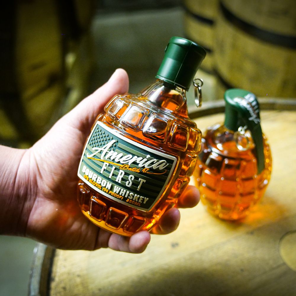 A hand holds an America First Grenade Straight Bourbon Whiskey bottle by America First, shaped like a grenade, with another in the background—ideal for patriotic gifting. Available as a 2 Pack.