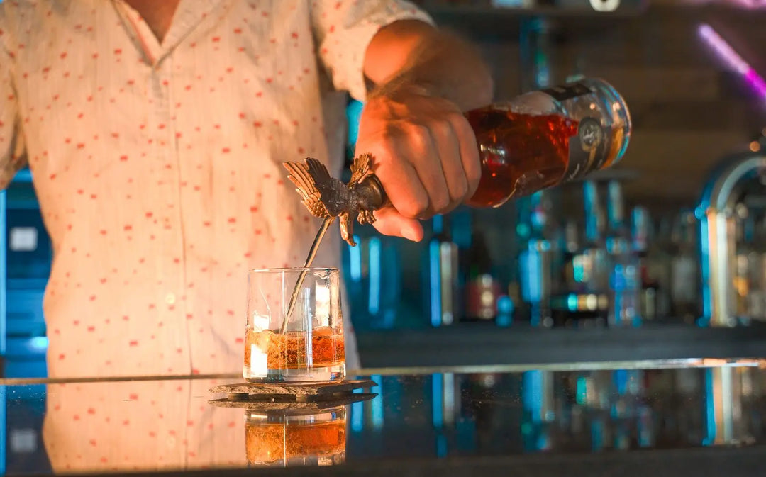 A bartender pours America First 250 Year Limited Edition Bourbon—a special 10-year-aged release—into a glass with ice from a bottle topped with an eagle-shaped stopper, set on a reflective bar counter.