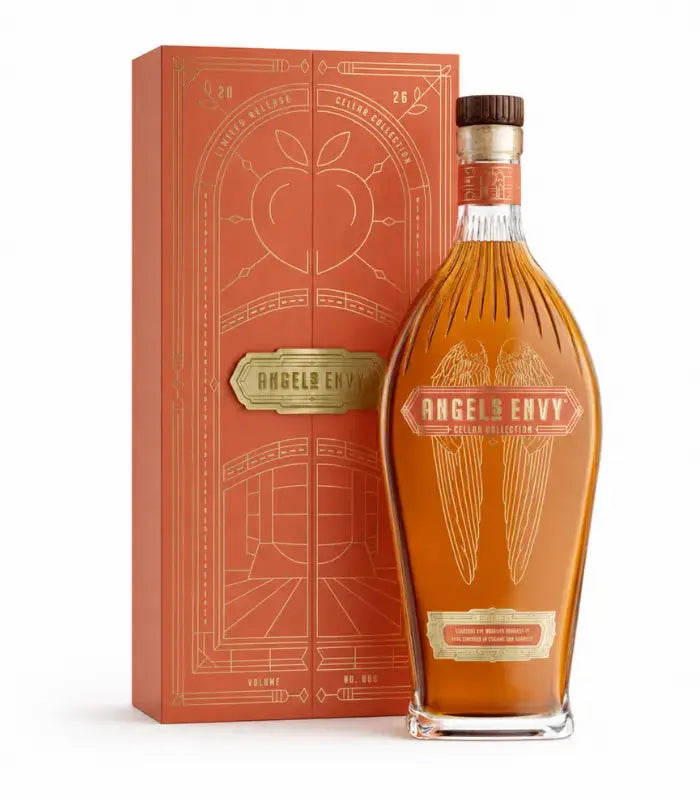 Standing before a vibrant orange and gold decorative box, a bottle of Angel’s Envy Cellar Collection Vol. 6 2026 Peach Brandy &amp; Toasted Oak Finished Rye Whiskey features elegant gold lettering and signature angel wing motifs. This limited release from Angel’s Envy is expertly crafted rye whiskey, uniquely finished in peach brandy and toasted oak casks for a distinctive richness of character.
