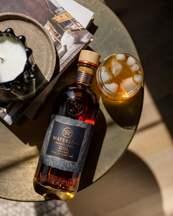 A bottle of Waterloo Barrel-Aged Gin by Waterloo Gin rests on a round table next to an iced cocktail, a candle, and a stack of books or magazines, all bathed in sunlight.