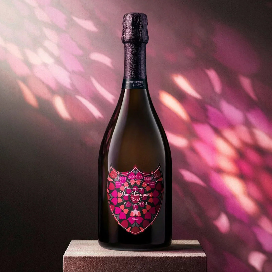 A bottle of Dom Pérignon Rosé Vintage 2010 Damien Hirst Limited Edition Champagne with a decorative, multi-colored floral label stands on a square stone pedestal against a pink and purple patterned background.