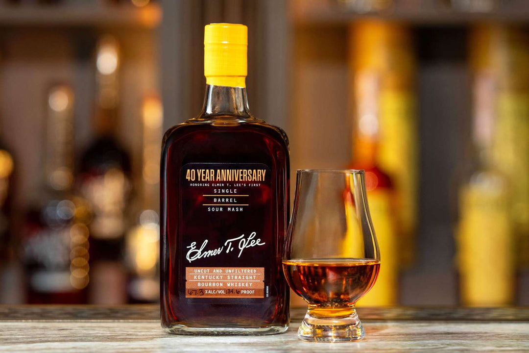A bottle of Elmer T. Lee 40 Year Anniversary Uncut &amp; Unfiltered Single Barrel Bourbon 127.8 Proof sits beside a whiskey glass of amber liquid on a wooden surface, with blurred bottles in the background.