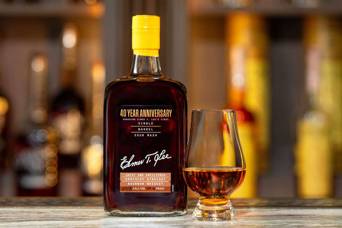 A bottle of Elmer T. Lee 40 Year Anniversary Uncut & Unfiltered Single Barrel Bourbon 127.8 Proof sits beside a whiskey glass of amber liquid on a wooden surface, with blurred bottles in the background.