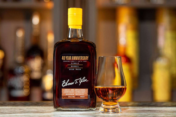 A bottle of Elmer T. Lee 40 Year Anniversary Uncut &amp; Unfiltered Single Barrel Bourbon 127.8 Proof sits beside a whiskey glass of amber liquid on a wooden surface, with blurred bottles in the background.