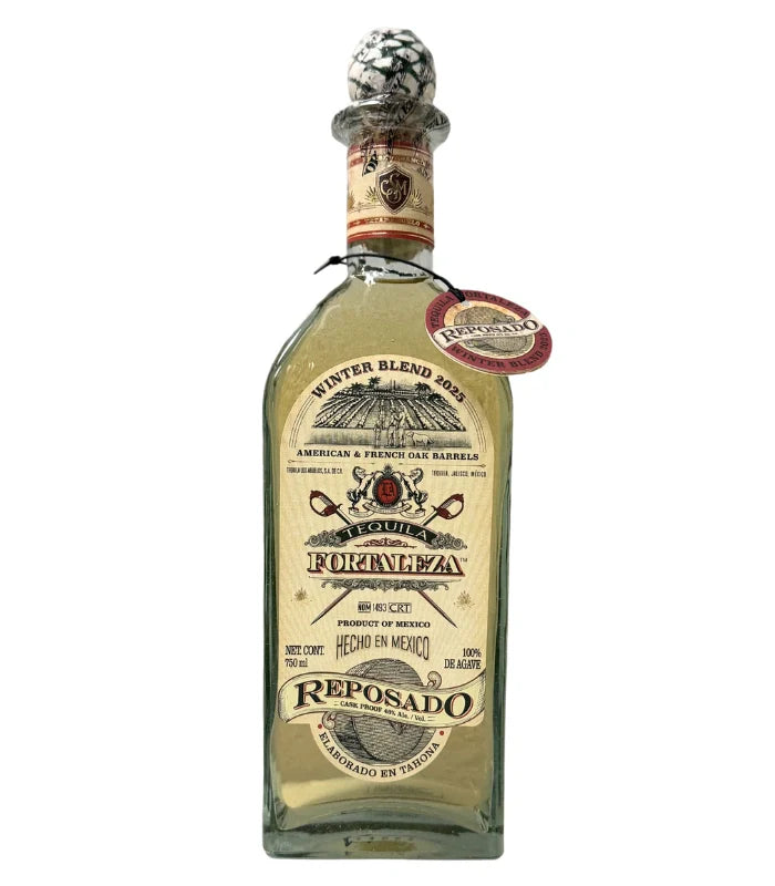 A clear glass bottle of Tequila Fortaleza Reposado Tequila Winter Blend 2025 with a beige label, decorative details, round agave-patterned stopper, and neck tag—an elegant limited edition presentation from Tequila Fortaleza.