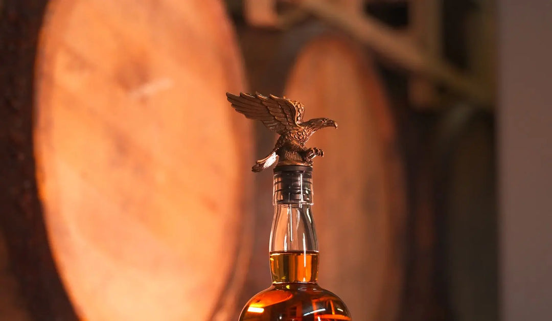 A close-up of the America First 250 Year Limited Edition Bourbon bottle by America First, featuring a bronze eagle stopper, with large wooden barrels blurred in the background.