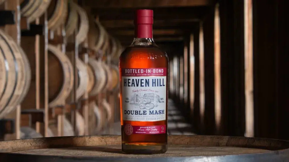 A bottle of Heaven Hill Double Mash Bottled-in-Bond Bourbon (Pre-Order) rests on a barrel inside a dimly lit rickhouse, with rows of wooden barrels stacked in the background.