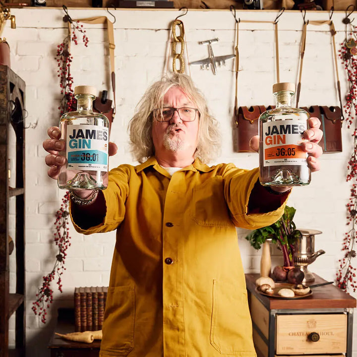 A gray-haired man in glasses and a yellow jacket stands indoors, holding two bottles of James Gin The Internationalist W/ Souvenir Gift Key Chain by James Gin US. Tools, hanging herbs, and vintage decor are visible in the background.