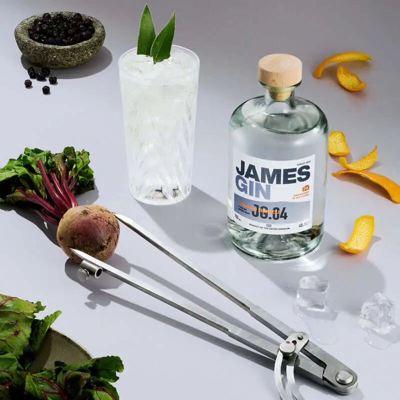 A bottle of James Gin London Drizzle 700mL 80 Proof with Souvenir Gift Key Chain, a crystal cocktail glass with mint, artisan botanicals, beetroot, orange peels, ice cubes, and spices all arranged on a white surface.