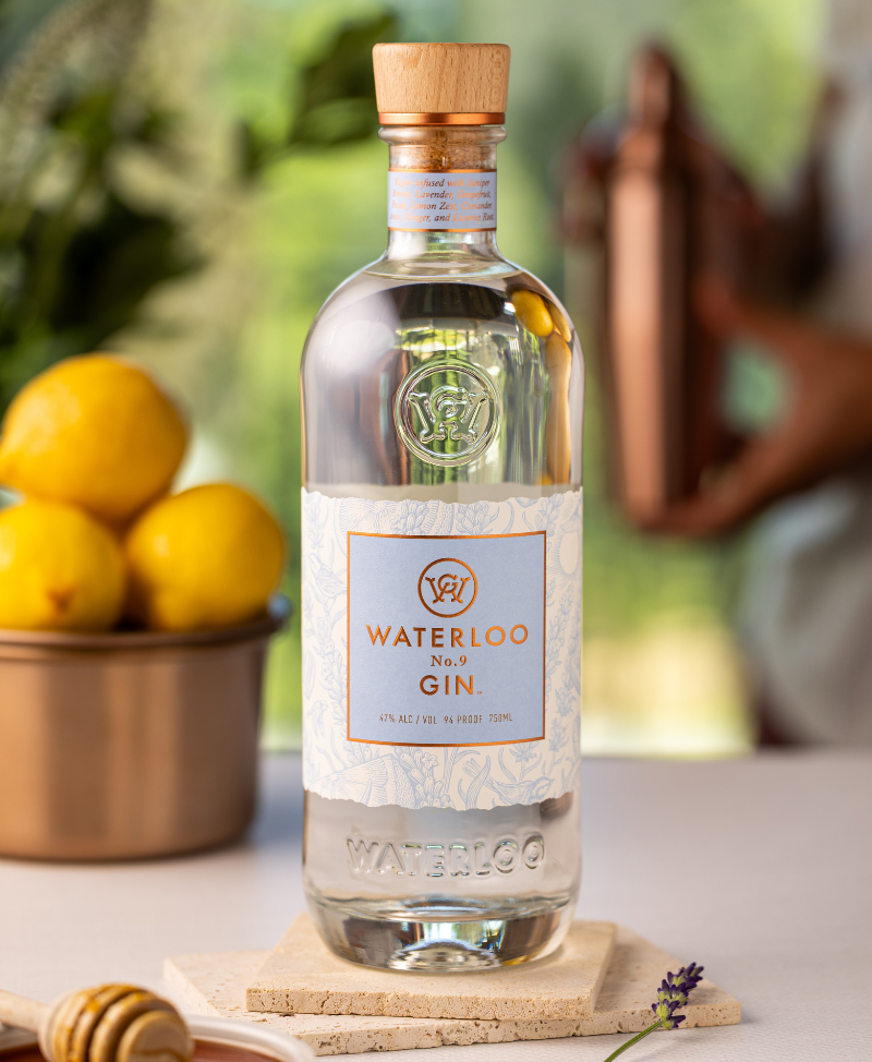 A clear bottle of Waterloo No.9 Gin by Waterloo Gin rests on a coaster. Behind it, lemons, a honey dipper, and someone holding a copper shaker create charm, with hints of greenery in the background.