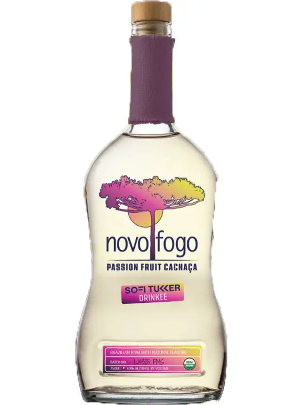 A clear glass bottle of Novo Fogo Passion Fruit Cachaça features a cork top and colorful tree artwork on the label, showcasing the brand’s vibrant Brazilian heritage with SOFI TUKKER DRINKEE text against a white background.