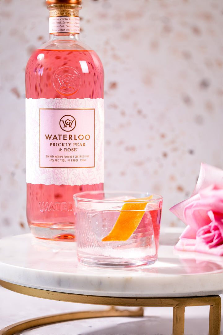A bottle of Waterloo Prickly Pear & Rose Gin by Waterloo Gin sits on a marble table beside a glass with clear liquid and an orange peel, displayed with a pink rose and a softly blurred background.