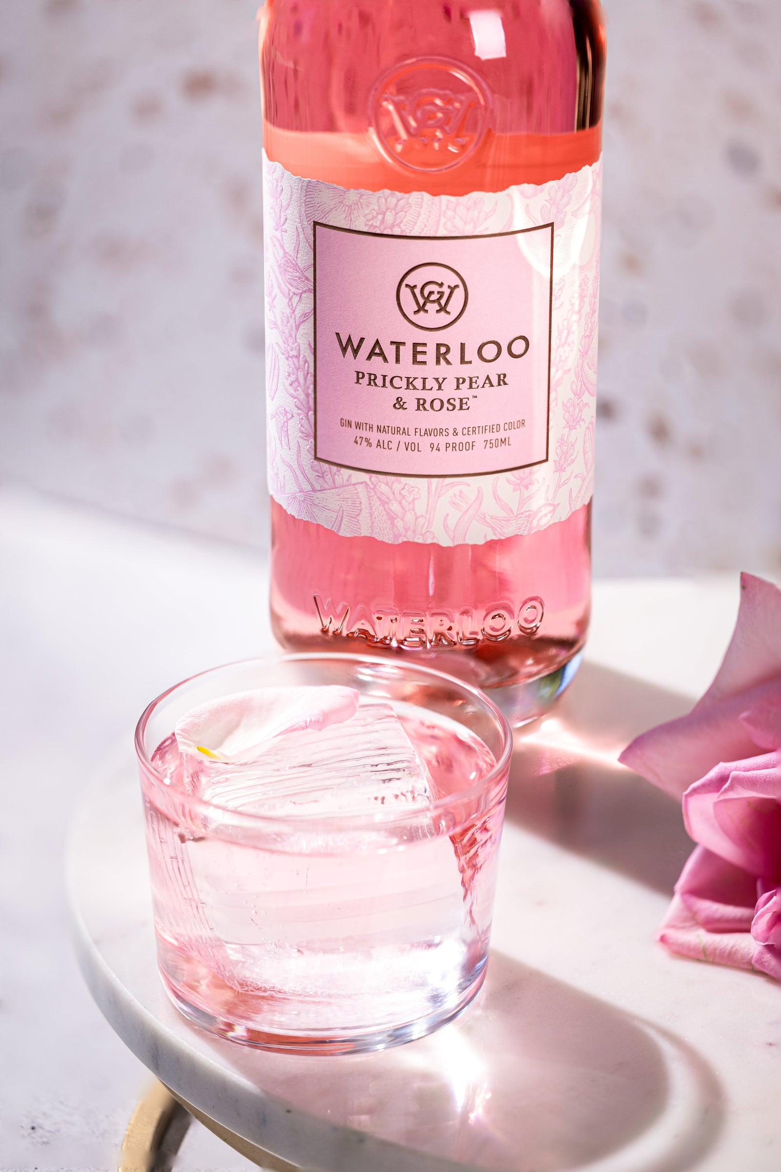 A bottle of Waterloo Gin's Prickly Pear & Rose Gin, its vibrant pink hue shining, stands behind a glass of ice and rose-tinted drink on white marble with a single pink rose petal, evoking the freshness of Texas Hill Country.