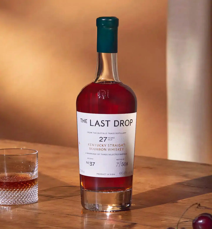 A bottle of The Last Drop Release No. 37 Buffalo Trace 27 Year Old Kentucky Straight Bourbon Whiskey 700ml, crafted by the acclaimed Buffalo Trace brand, stands on a wooden surface next to a glass of whiskey with ice, both illuminated by warm sunlight.