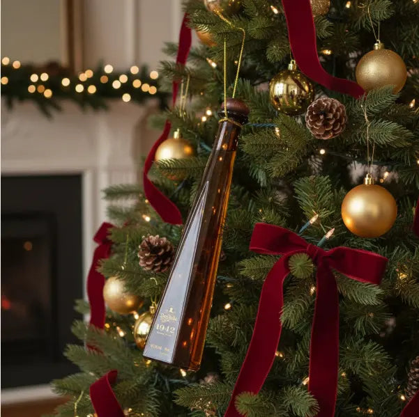 A Christmas tree adorned with red ribbons, gold ornaments, pinecones, and Don Julio 1942 Tequila Añejo 50ml Ornament 10-Pack glows by the fireplace—an elegant holiday touch from Don Julio Tequila for festive gifting.