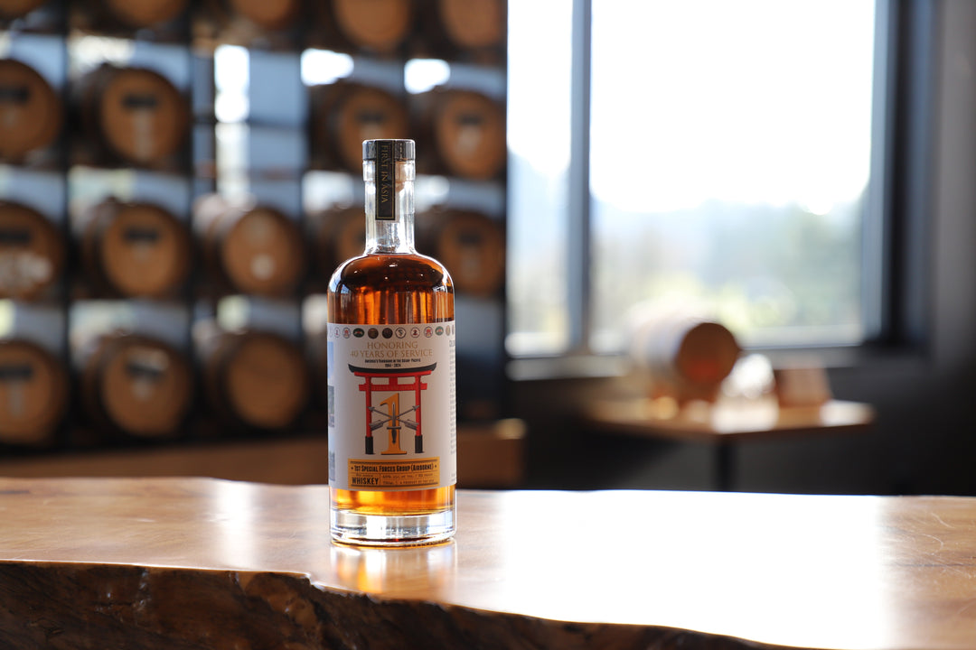 A bottle of Heritage Distilling’s 1st Special Forces Group Whiskey (8th Edition), a Bourbon Rye blend with a red torii gate label, stands on a wooden table in the distillery, barrels and a window blurred in the background.