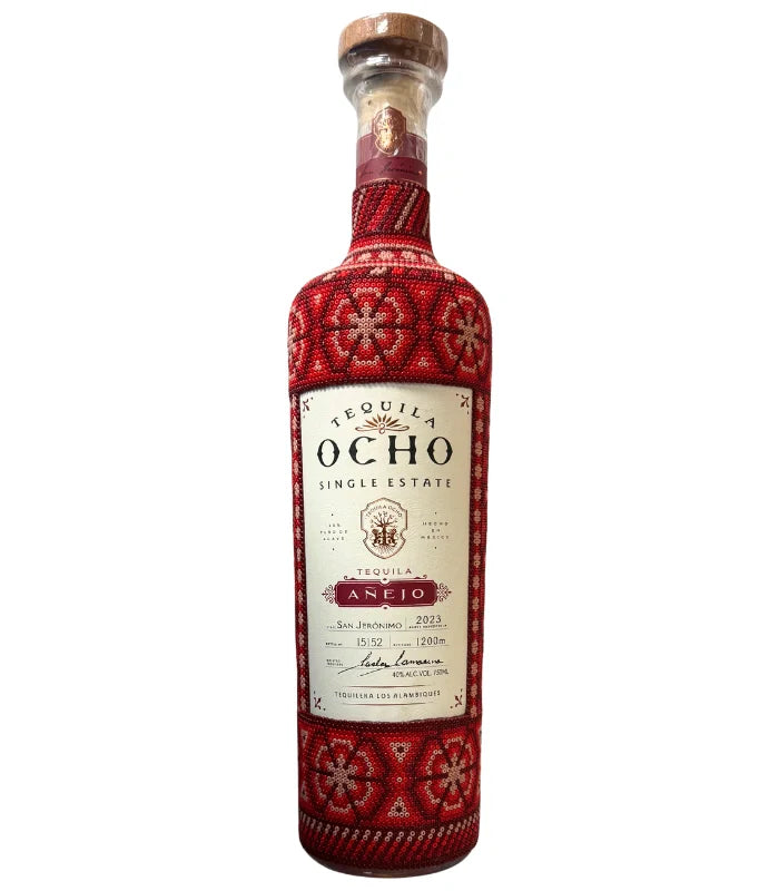 A bottle of Tequila Ocho Añejo Huichol Art Edition by Ocho Tequila, featuring a beige label and a red beaded cover with detailed floral and geometric patterns, finished with a cork.