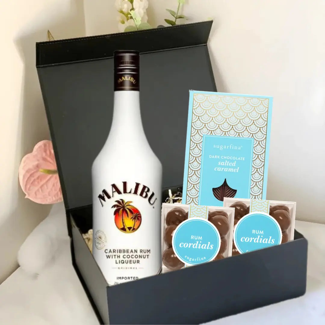 The Premium Rum Gift Box by Main Street Liquor features Malibu Caribbean Rum with Coconut Liqueur, Sugarfina dark chocolate sea salt caramels, and two packs of Sugarfina Rum Cordials. Pink flowers provide an elegant background touch.