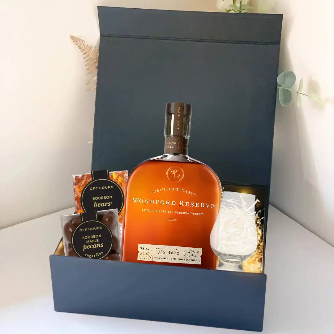 The Premium Bourbon Gift Box by Main Street Liquor features Woodford Reserve whiskey, a glass, bourbon bears gummies, and bourbon pecans—all elegantly arranged against a plain background.