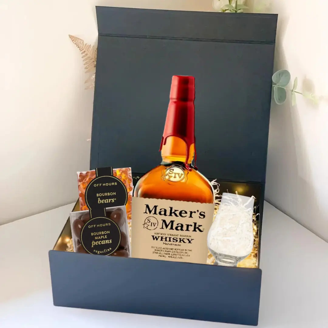 The Premium Bourbon Gift Box from Main Street Liquor features Maker’s Mark bourbon, a whiskey glass, pecans, bourbon bears, and a snack bar arranged with decorative lights in a sleek black gift box.