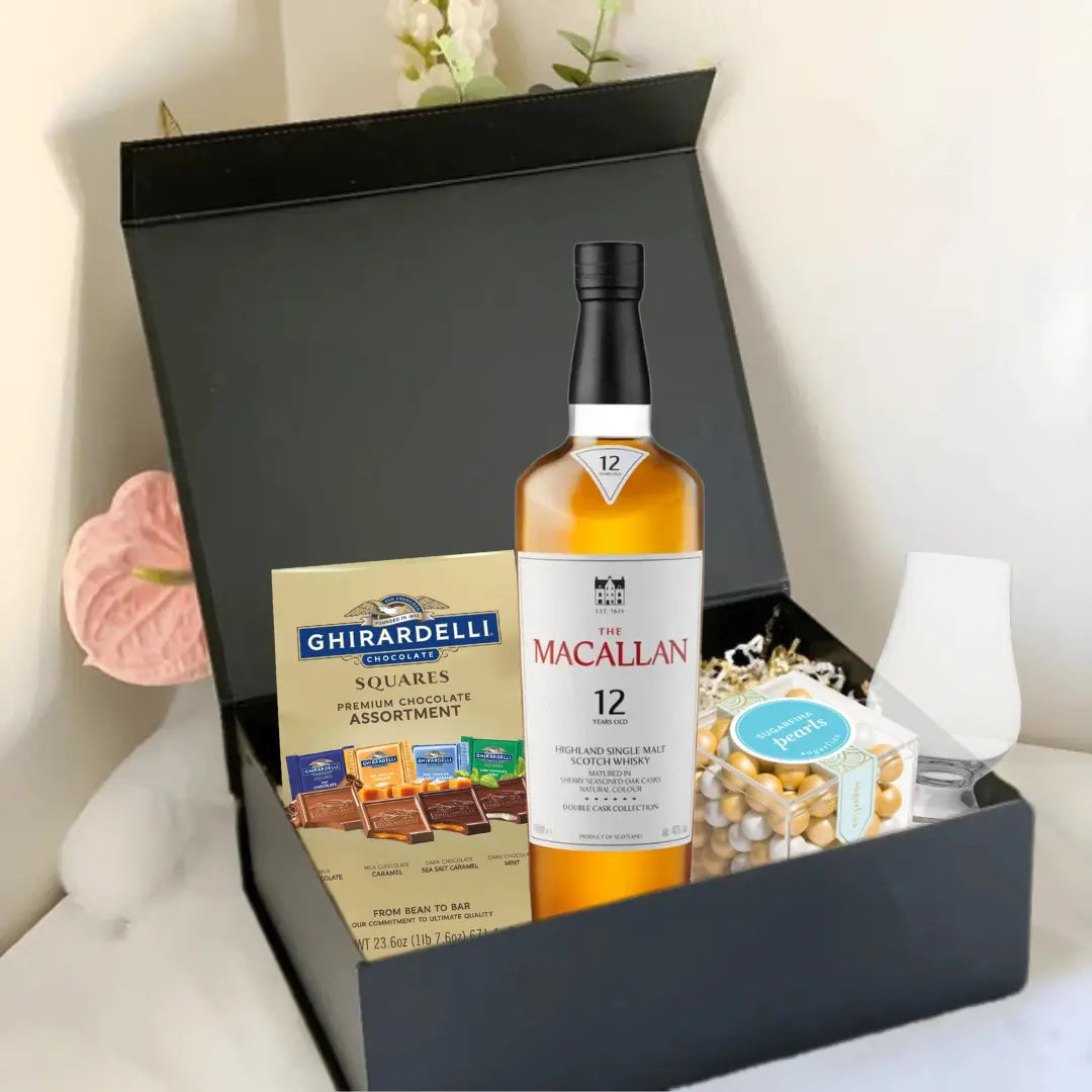 The Premium Scotch Gift Box by Main Street Liquor includes Macallan 12 whisky, Ghirardelli chocolate squares, a clear glass, and Ferrero Rocher chocolates, all in a black box set against a white surface with pink flowers.