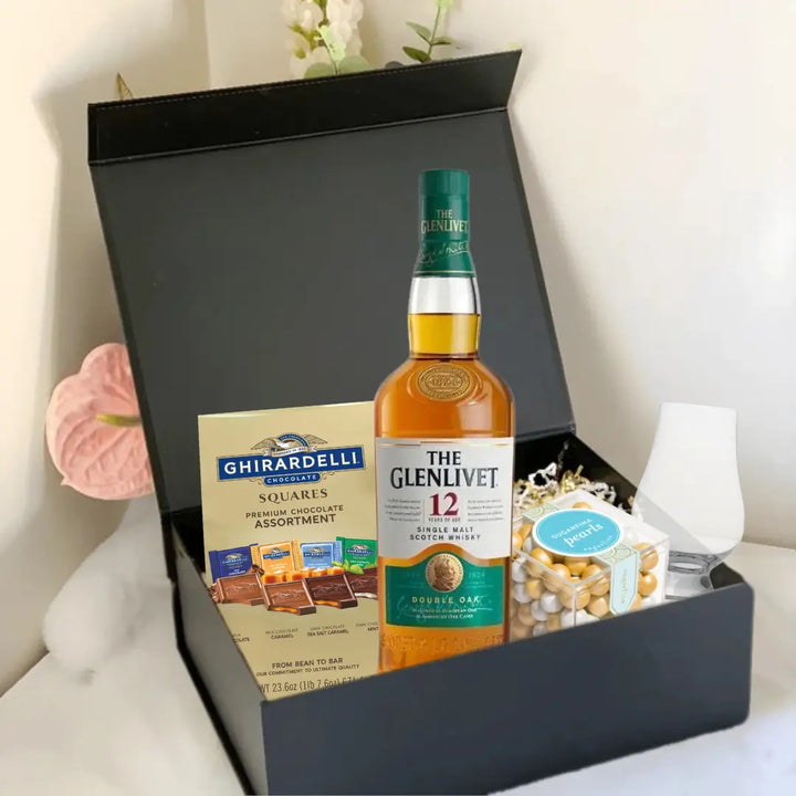 The Premium Scotch Gift Box by Main Street Liquor includes The Glenlivet 12-Year-Old Scotch, Ghirardelli chocolates, a glass, and mixed nuts, elegantly arranged with floral accents.