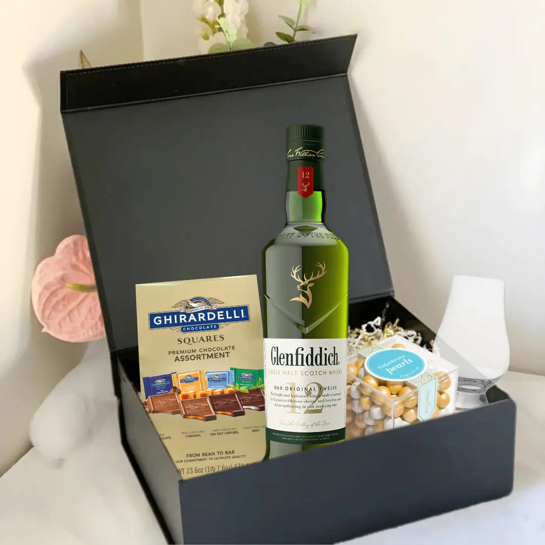 The Premium Scotch Gift Box by Main Street Liquor features Glenfiddich 12 Year Old whisky, Ghirardelli chocolate squares, a glass, and mixed nuts, all elegantly arranged with decorative flowers in the background.
