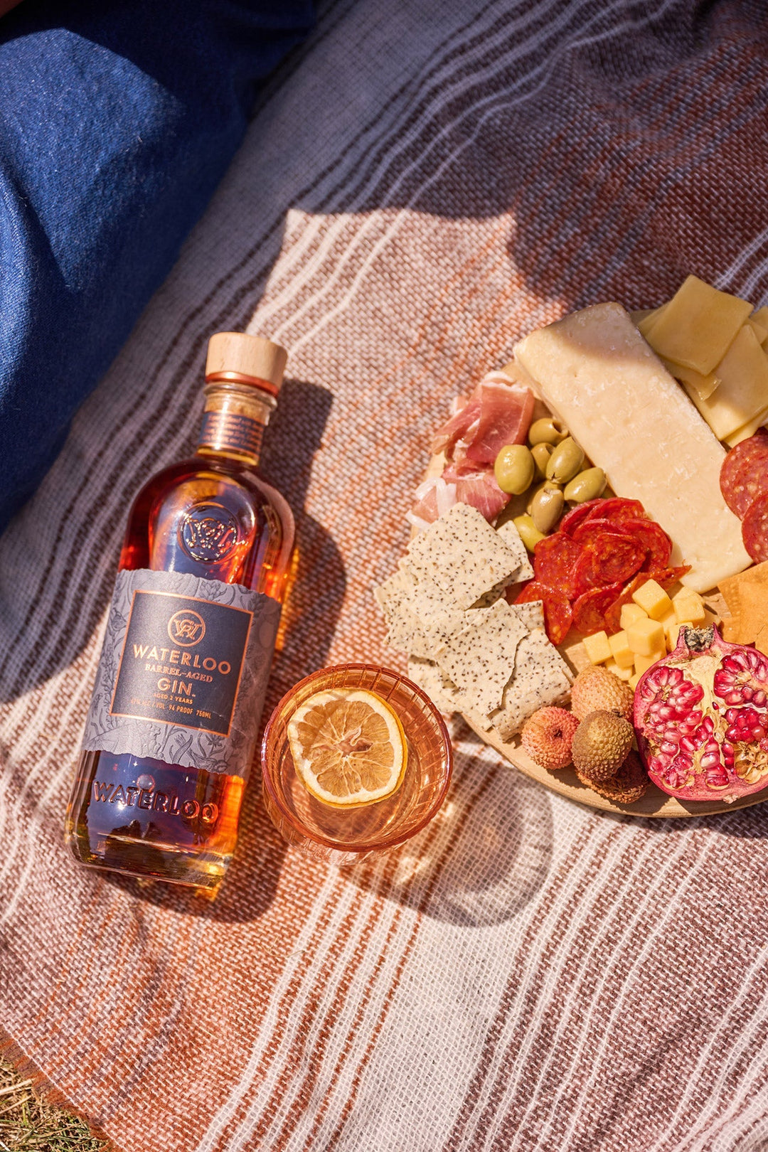 A bottle of Waterloo Barrel-Aged Gin by Waterloo Gin, a glass with a dried citrus slice, and a plate of assorted cheeses, meats, crackers, olives, and fruit are set out on a striped picnic blanket.