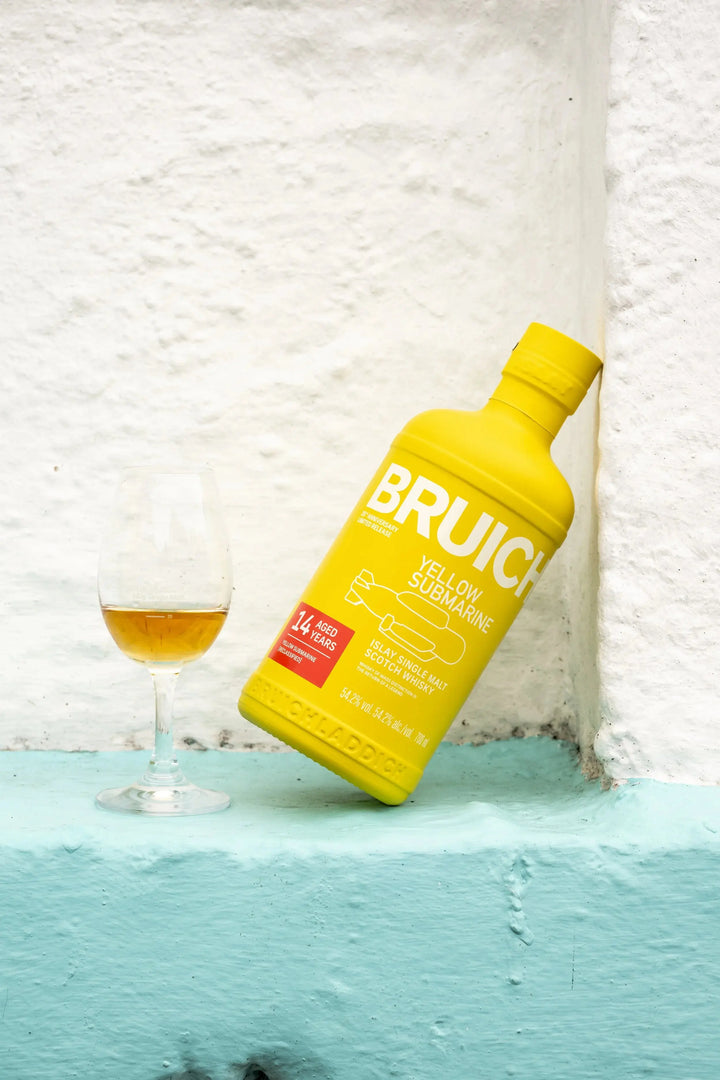 A yellow bottle of Bruichladdich Yellow Submarine III 14 Year Old Islay Single Malt Scotch Whisky leans against a white wall, beside a glass of amber liquid on a light blue ledge.