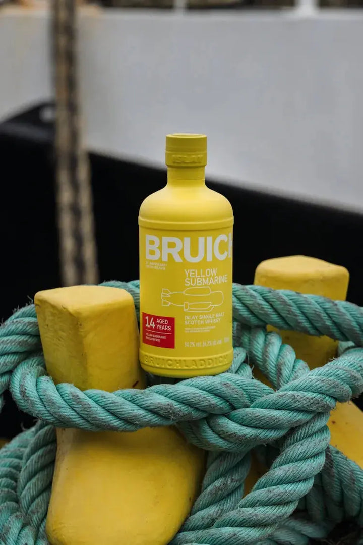 A yellow bottle of Bruichladdich Yellow Submarine III 14 Year Old Islay Single Malt Scotch Whisky sits on yellow bollards with teal rope, with an out-of-focus nautical background.