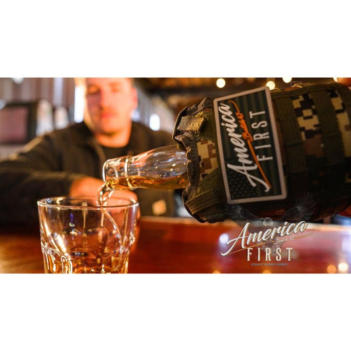 A close-up of an America First Straight Bourbon Whiskey 2 Pack bottle in a camouflage sleeve pouring bourbon into a glass, with a blurred man at the bar in the background.