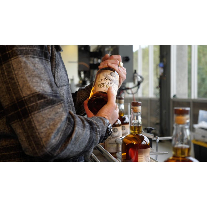 Wearing a plaid jacket, a person holds an America First Straight Bourbon Whiskey 2 Pack at a bar, with other America First bottles highlighting American craftsmanship on the counter by large windows.