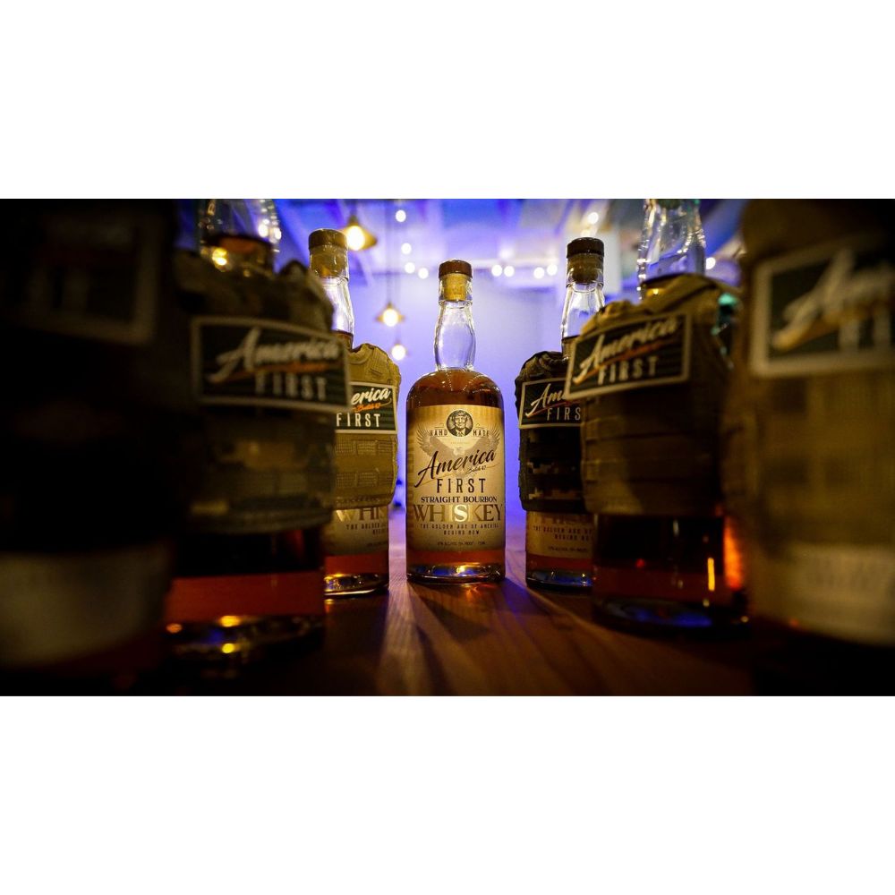 An America First Straight Bourbon Whiskey 3 Pack sits on a wooden table, surrounded by other bottles from this limited-edition set under warm lighting and glowing string lights, creating an inviting, nostalgic mood.