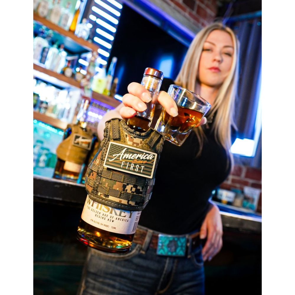 A woman in a bar holds a glass of whiskey and extends an America First Straight Bourbon Whiskey 2 Pack (in camouflage holder) toward the camera, with shelves of bottles and a brick wall in the background.