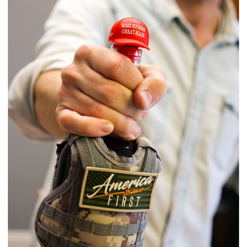 A person in a light-colored, long-sleeved shirt holds an America First Straight Bourbon Whiskey 6 Pack bottle with a red cap reading MAKE BOURBON GREAT AGAIN, featuring a limited-edition camouflage sleeve and America First patch.
