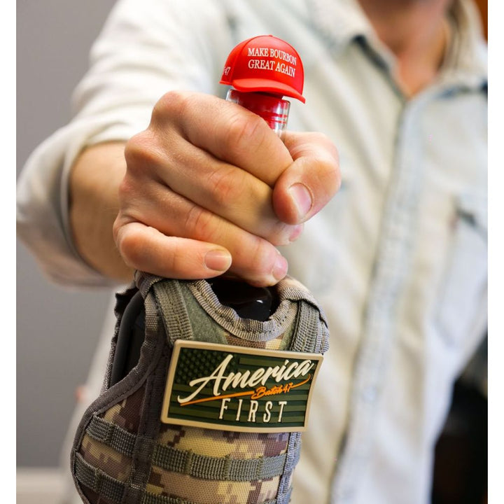 A person holds an America First Straight Bourbon Whiskey 3 Pack bottle with a red cap reading “MAKE BOURBON GREAT AGAIN,” in a camouflage sleeve featuring an “America FIRST” patch from the America First brand.