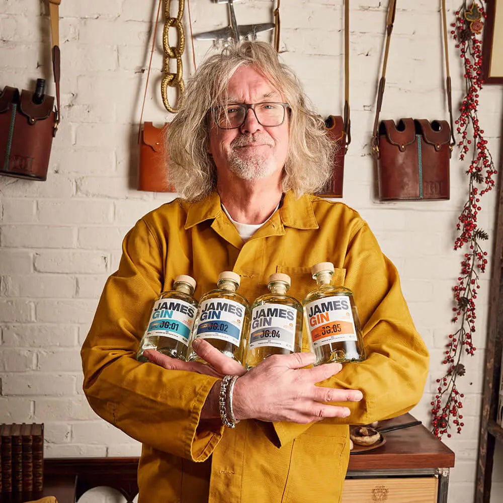 A man with shoulder-length gray hair and glasses, in a mustard yellow jacket, stands before a brick wall holding the James Gin The Mixologist's Cocktail Kit w/ Souvenir Gift Key Chain from James Gin US; tools and leather bags hang behind him.
