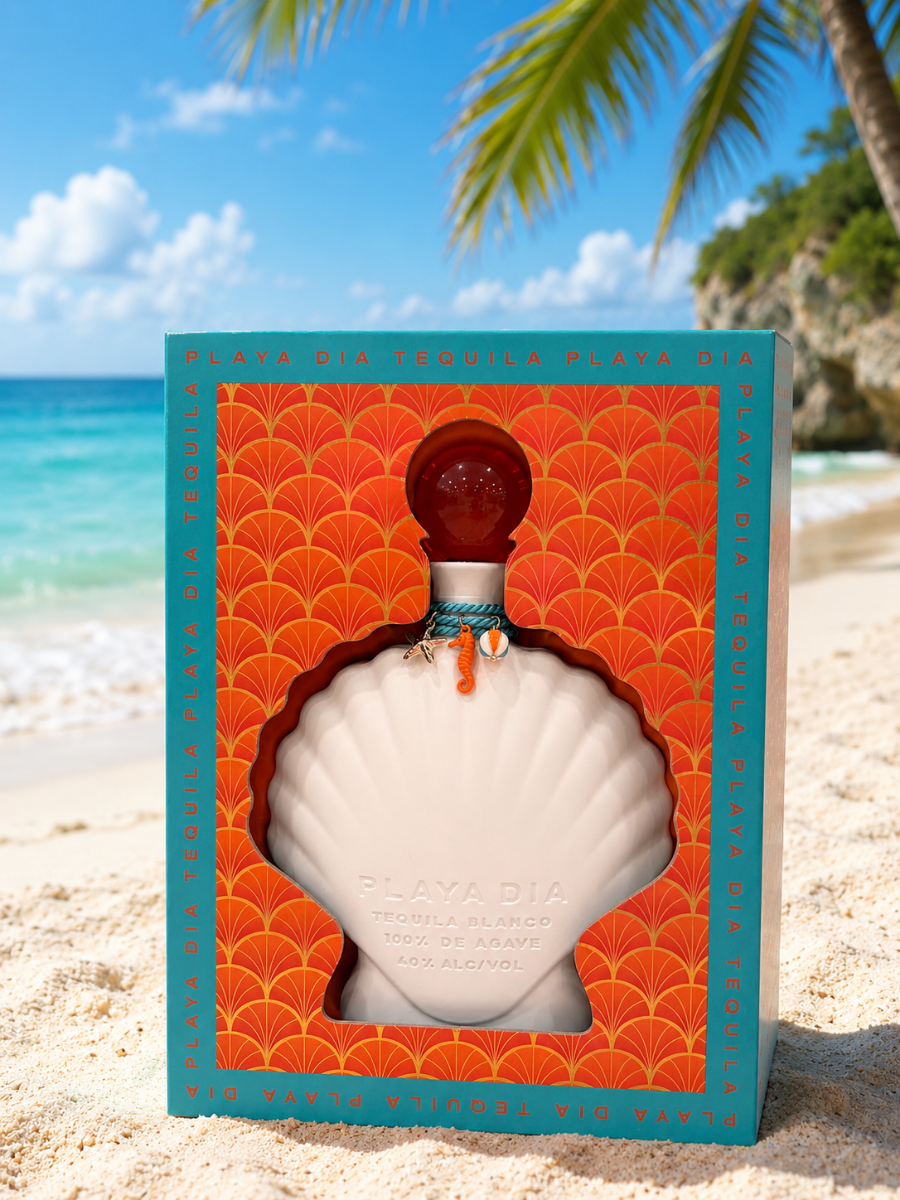 A bottle of PLAYA DIA Blanco Tequila, crafted from 100% Blue Weber agave and entirely additive-free, rests in a shell-shaped box on a sandy beach with a palm tree and clear blue ocean in the background. The orange packaging with teal accents beautifully complements this exceptional spirit from Playa Dia Blanco Tequila.