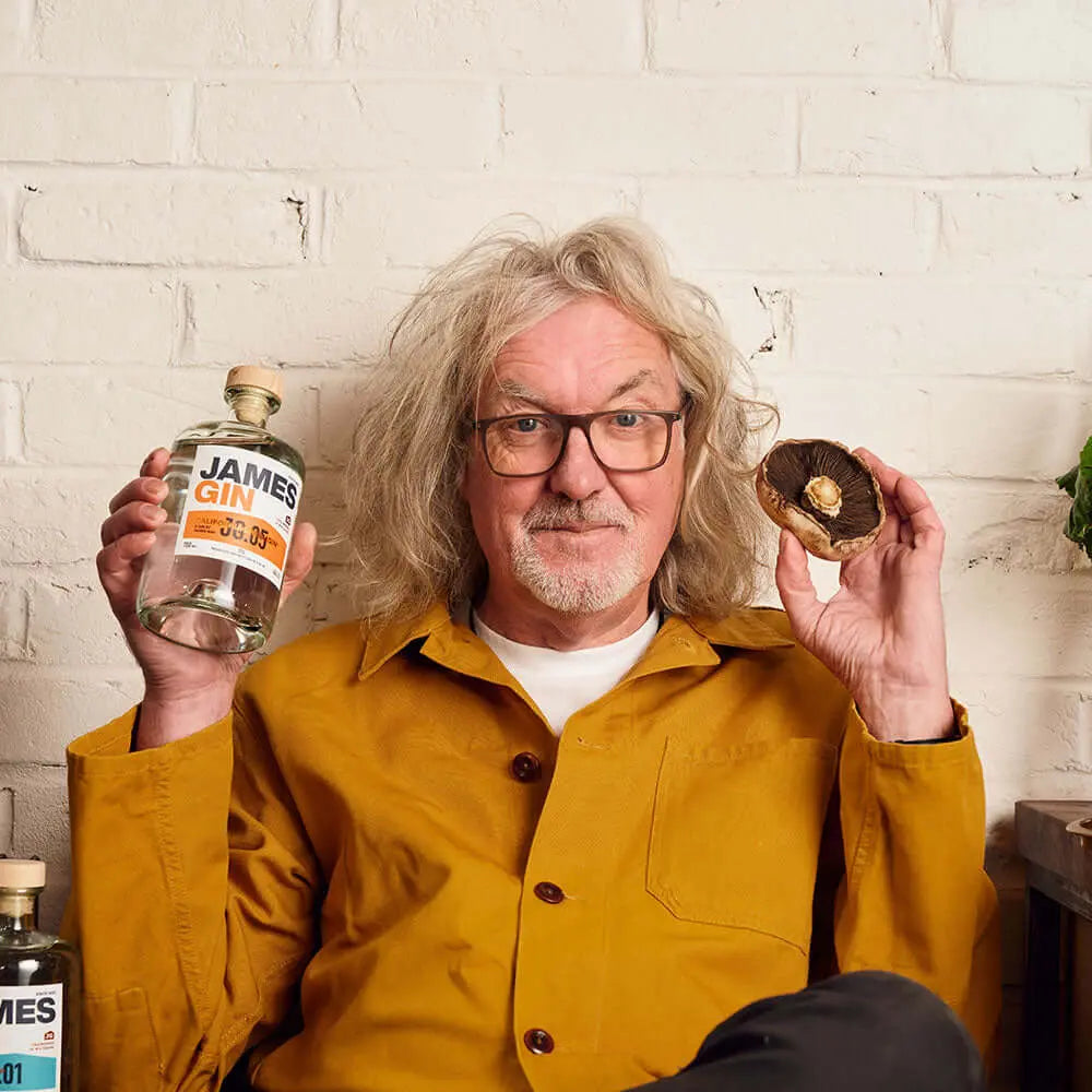 A man with gray, curly hair and glasses, in a mustard jacket, sits before a white brick wall holding a James Gin US The Internationalist bottle with souvenir gift key chain in one hand and a large portobello mushroom in the other.