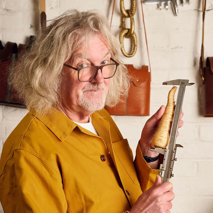 A man with long gray hair and glasses, in a mustard-yellow shirt, smiles while measuring a parsnip—perhaps testing ingredients for James Gin Navy Strength 700mL 114 Proof. Leather bags and tools hang on a white brick wall behind him.
