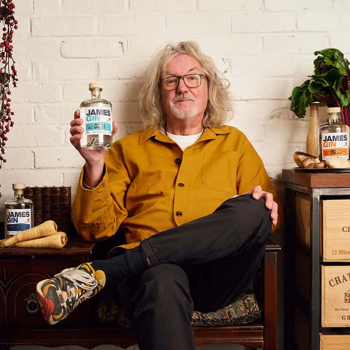 A man with shoulder-length gray hair, glasses, and a yellow jacket sits cross-legged, holding a James Gin Asian Parsnip 700mL 80 Proof with Souvenir Gift Key Chain. Other James Gin bottles and wooden crates rest nearby against a white brick wall.