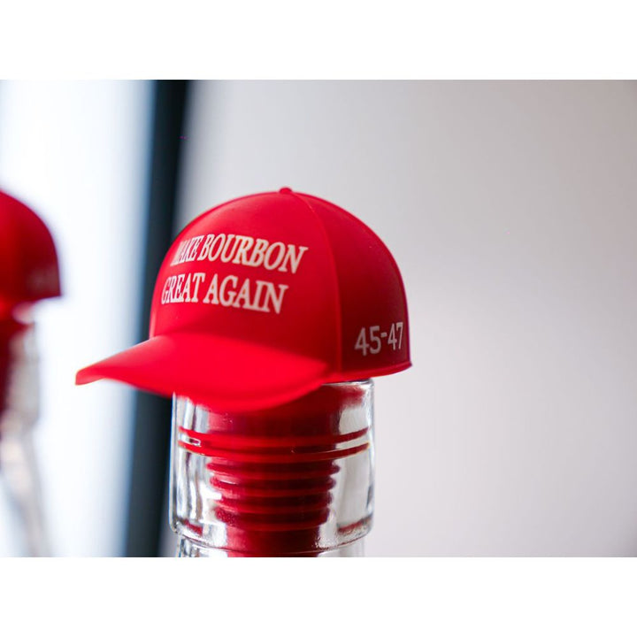 A red bottle cap shaped like a baseball hat with "MAKE BOURBON GREAT AGAIN" and "45-47" decorates the America First Straight Bourbon Whiskey 2 Pack, showcasing the brand’s dedication to American craftsmanship.